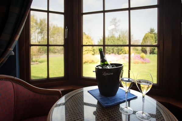 Una habitación con vistas amplias a través de ventanas grandes, una mesa con una botella de champán y dos copas, y una silla roja con estampado.