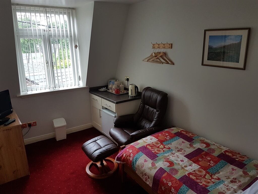 Une chambre avec un lit, un bureau, une chaise, une fenêtre avec des stores, une petite poubelle blanche et un tableau encadré au mur.