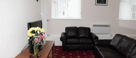 A living room with a black leather sofa, a wooden side table with a vase of flowers, and a patterned carpet.