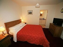 Un dormitorio con cama, lámpara de noche, cómoda, televisor y un pequeño cuadro en la pared.