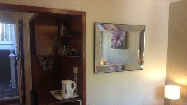 Habitación de hotel con cama, mesita de noche con lámpara, estante de madera con tetera y vasos, espejo y puerta que da al exterior.