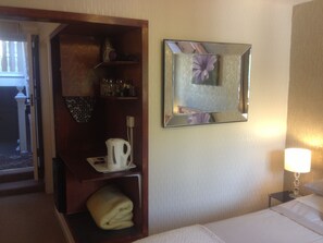 A hotel room with a bed, a nightstand with a lamp, a wooden shelf with a kettle and glasses, a mirror, and a door leading outside. - Cameron House Hotel (Oban)