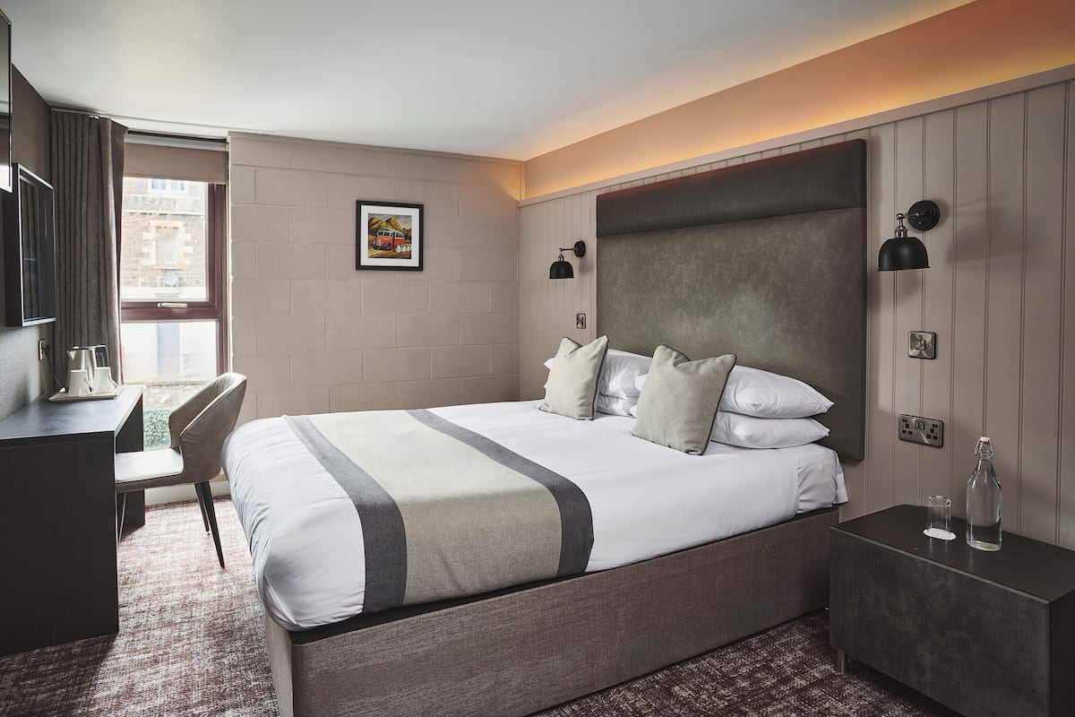 A hotel room with a bed, bedside table, desk, chair, and a window with curtains.