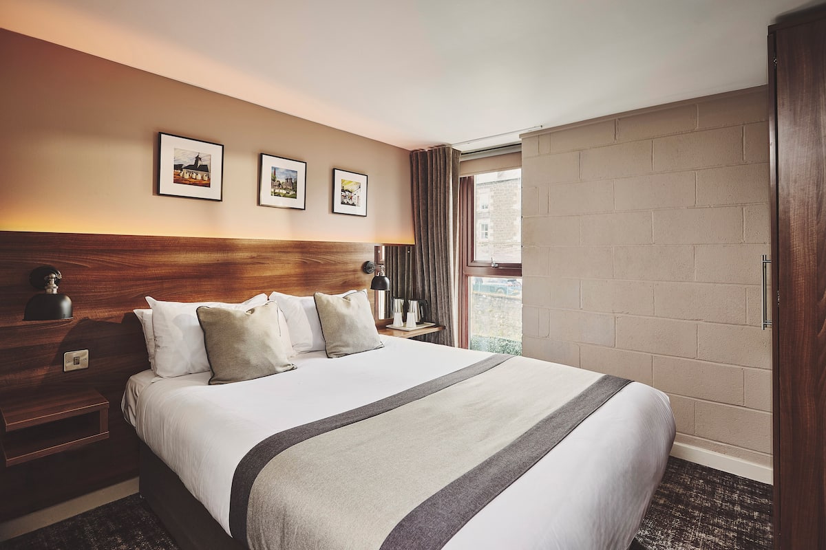 A hotel room with a large bed, a wooden headboard, a nightstand with a lamp, and three framed pictures on the wall.
