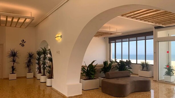 A spacious lobby with a curved sofa, potted plants, and a large window offering a view of the sea.