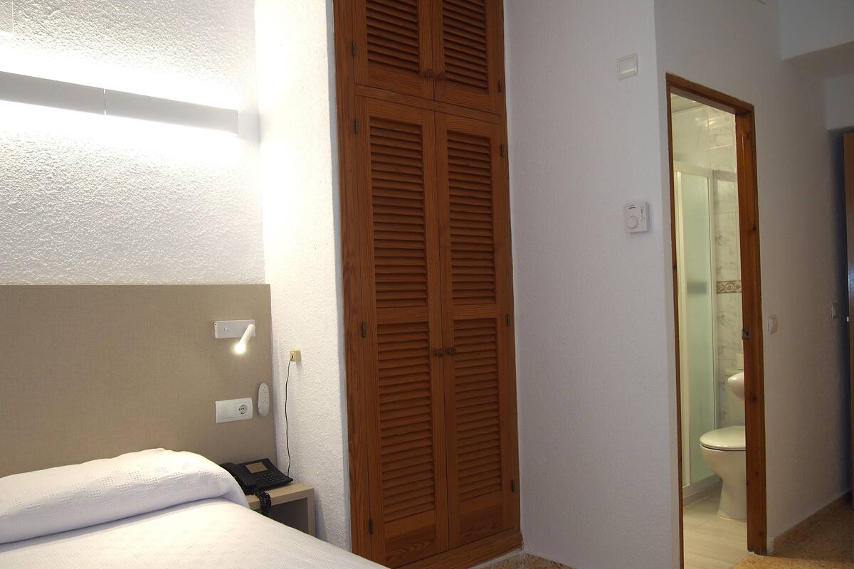 Ein Hotelzimmer mit einem Bett, einem Holzschrank, einem Nachttisch mit Telefon und einem durch eine offene Tür einsehbaren Badezimmer.