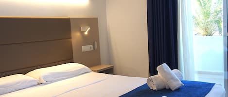 A hotel room with a bed, a nightstand, a wall-mounted light, and a view of greenery outside.