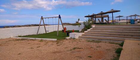 A beachside area with a swing set, steps leading to a covered seating area, and a view of the sea.