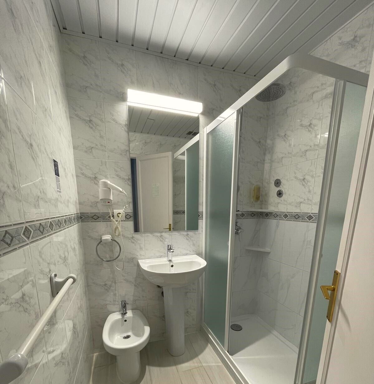 A bathroom with a marble floor, a pedestal sink, a bidet, a handheld showerhead, and a glass-enclosed shower.