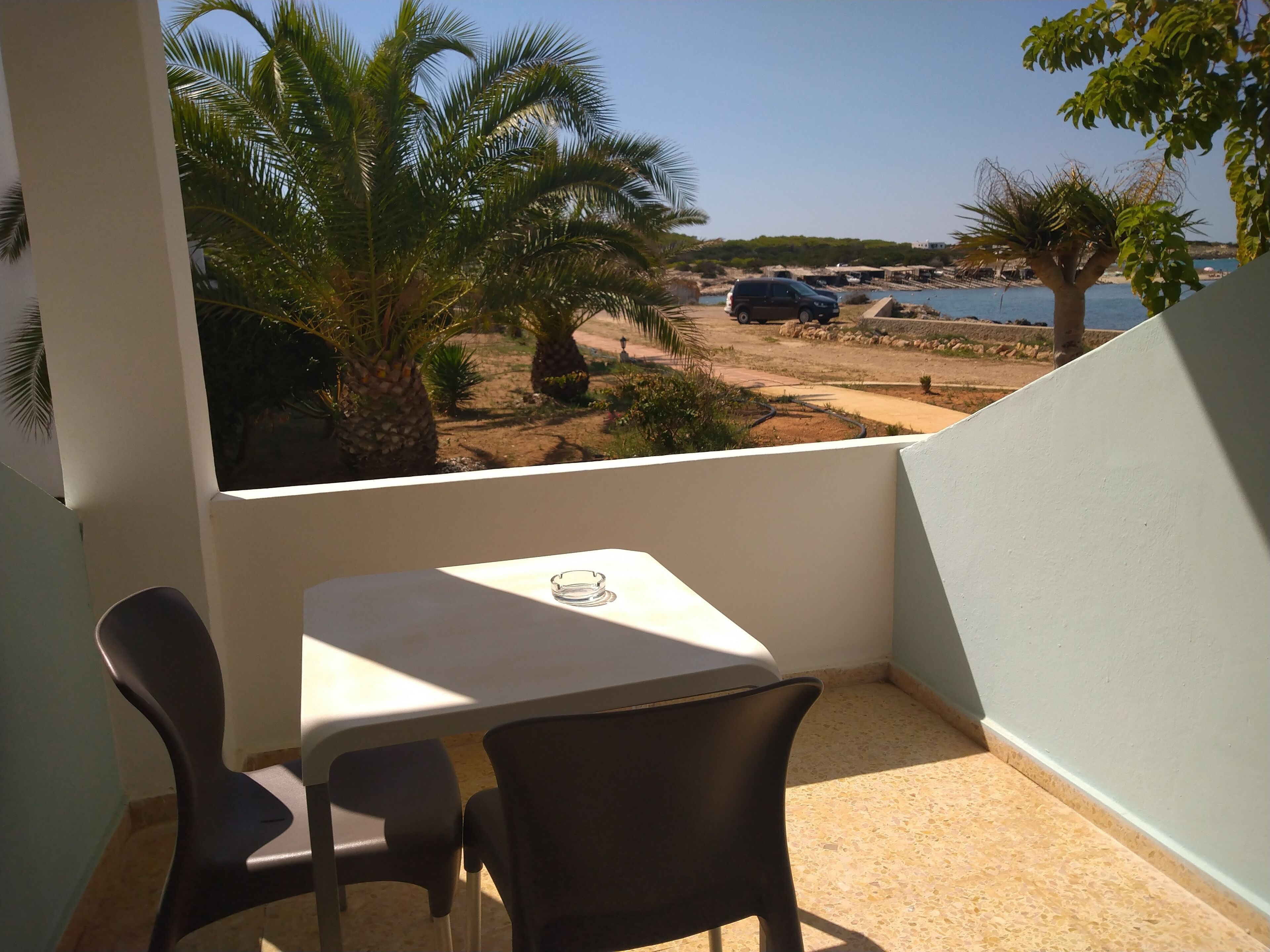 Ein Balkon mit Tisch und Stühlen, mit Blick auf einen Strand mit Palmen und einem geparkten Auto.