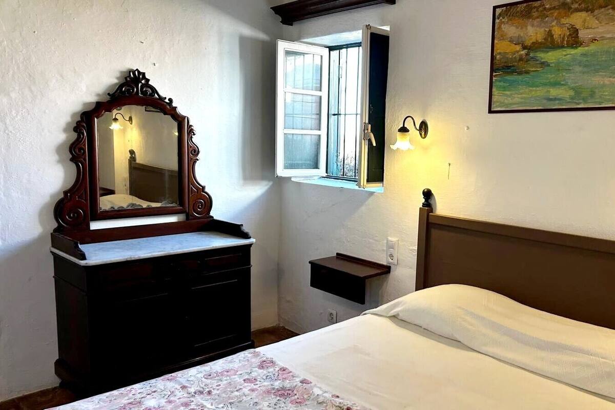 A bedroom with a wooden dresser, a bed with white bedding, a nightstand, a wall-mounted lamp, and a painting.