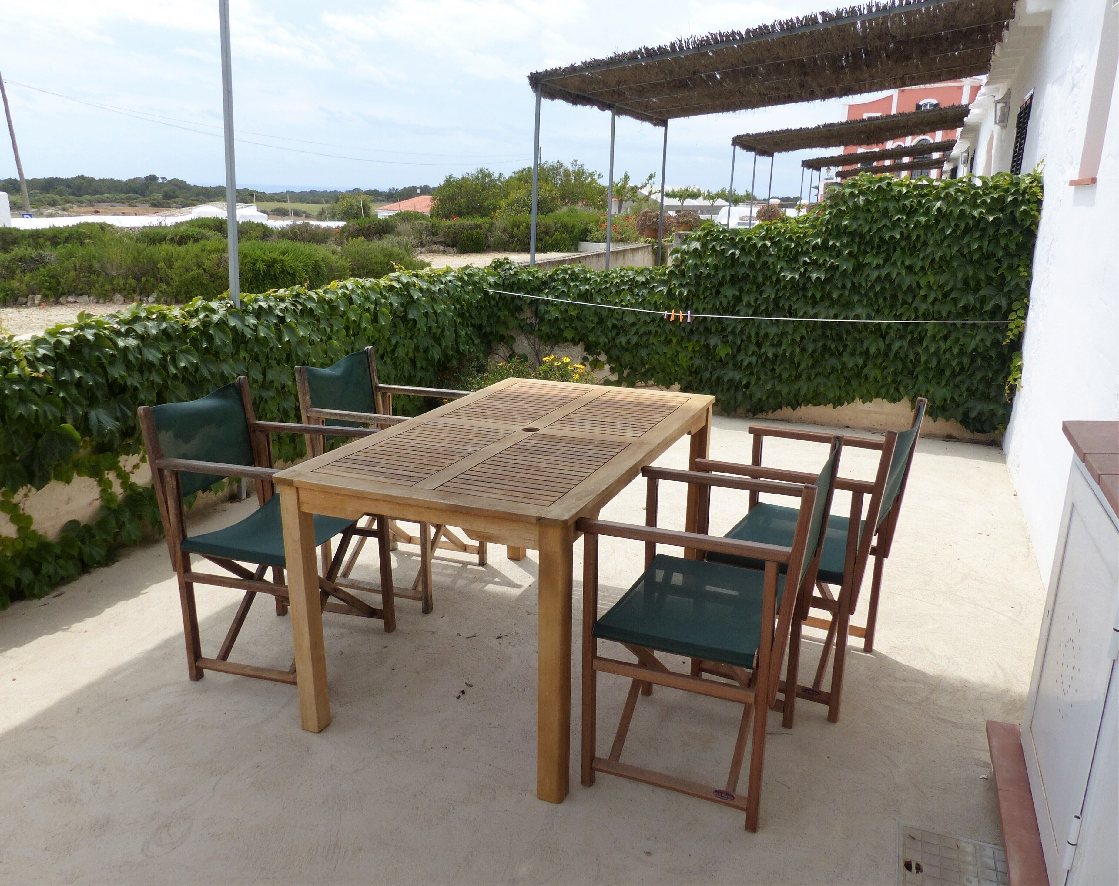 An outdoor patio with a wooden table and chairs, surrounded by greenery and a view of buildings in the distance.