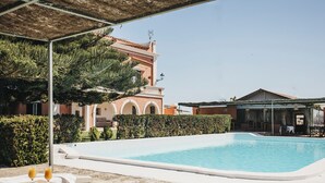 Un área de piscina con sillones y una mesa, un edificio con arcos y un cielo despejado.