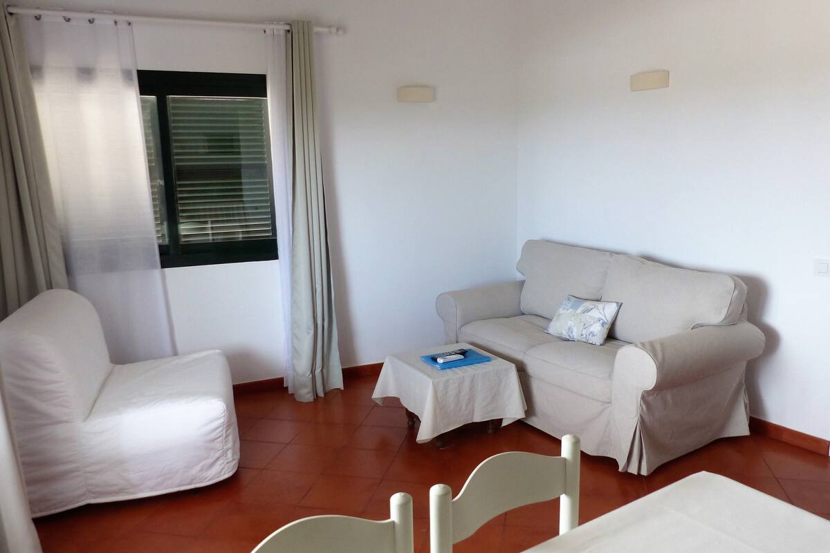 A room with a white sofa, a white chair, a small table with a remote control, and a window with curtains.