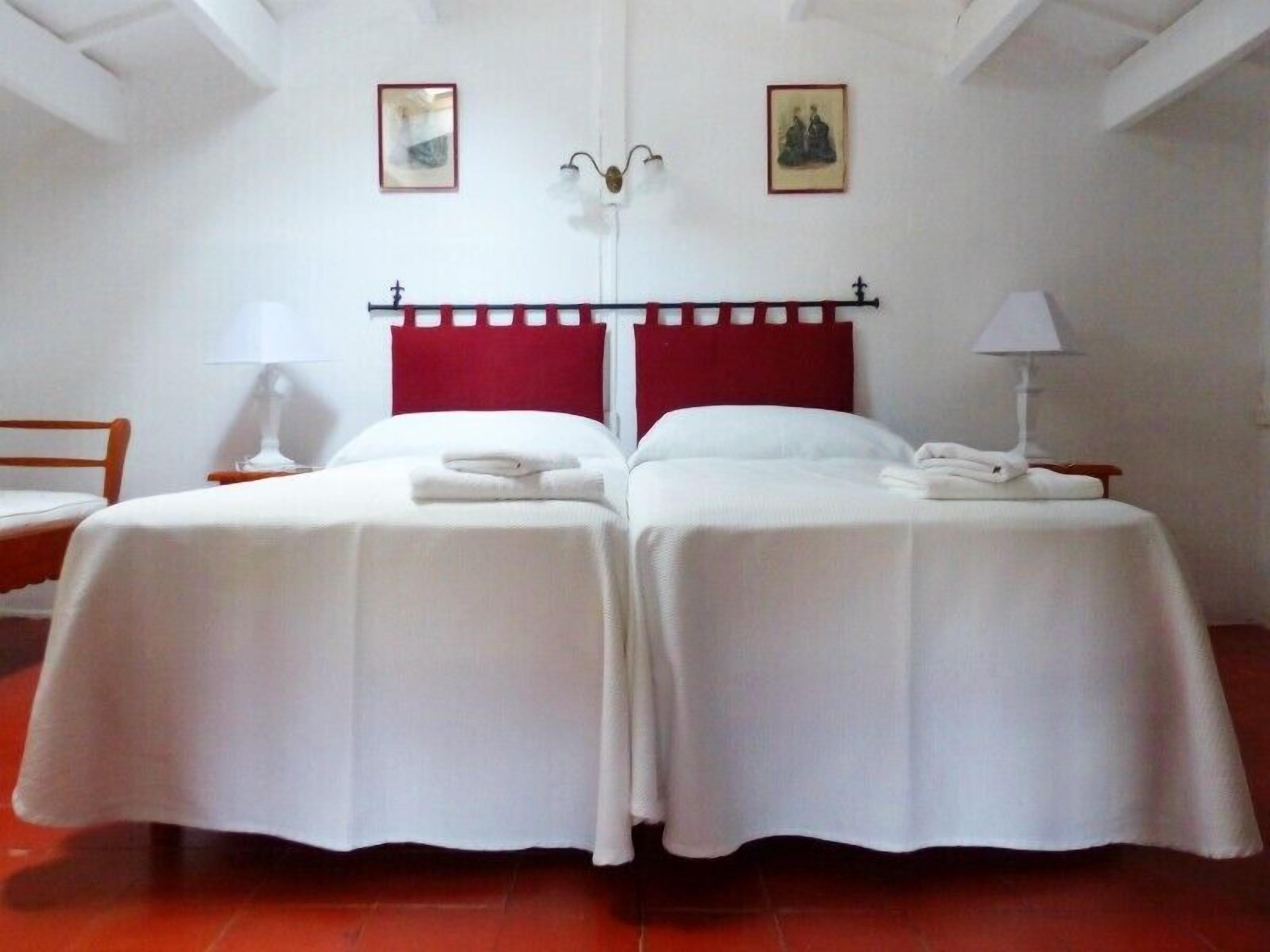 A double room with two beds, a red headboard, white bedding, a nightstand with a lamp, and two framed pictures on the wall.