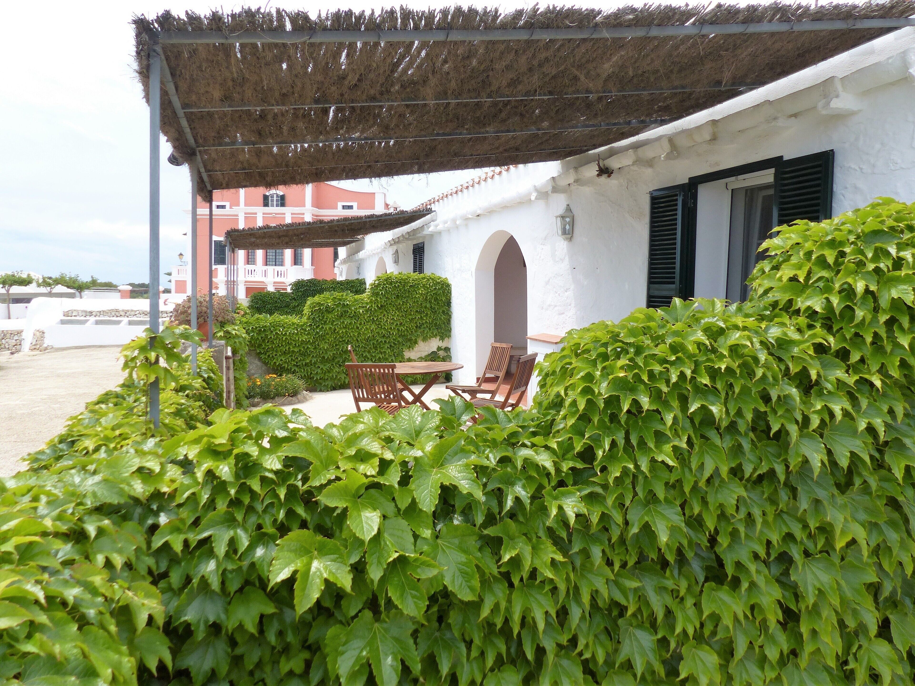 Un edificio blanco con techo de paja, un patio, una mesa y sillas, y un seto en primer plano.