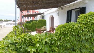 Un edificio blanco con techo de paja, un patio, una mesa y sillas, y un seto en primer plano.