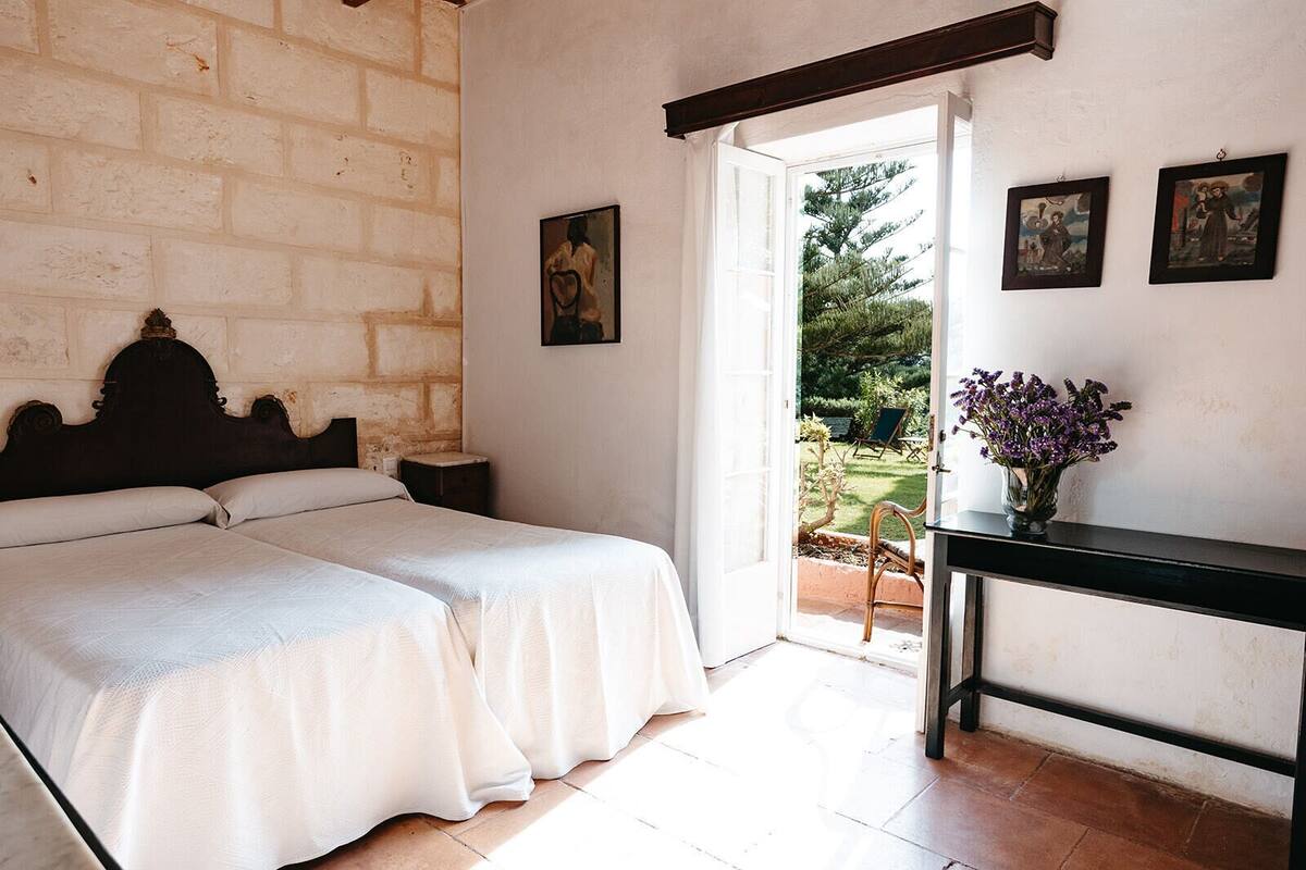 A bedroom with a double bed, a nightstand, a desk with a vase of flowers, and a view of the garden through an open door.