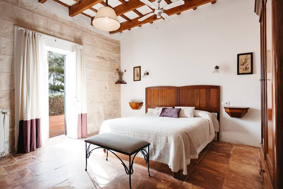 A bedroom with a wooden headboard, a bedside table, a bench, and a view of the outdoors through a glass door.