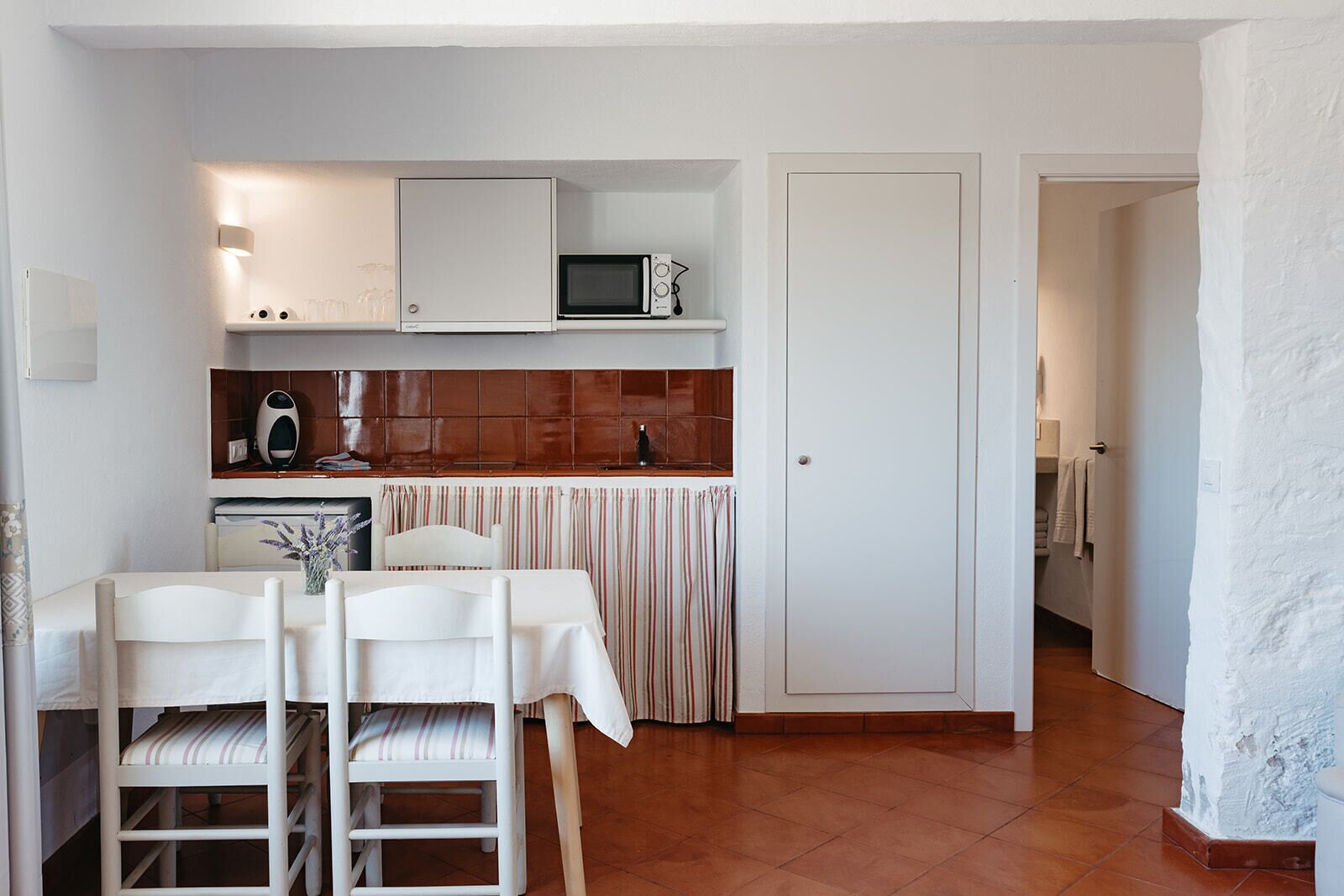 A compact kitchen with white cabinets, a microwave, and a small refrigerator. There is a dining table with chairs and a striped tablecloth.