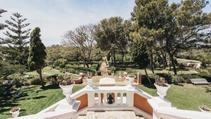 Un sendero de jardín que lleva a una estructura blanca con una fuente, rodeado de árboles y plantas en macetas.
