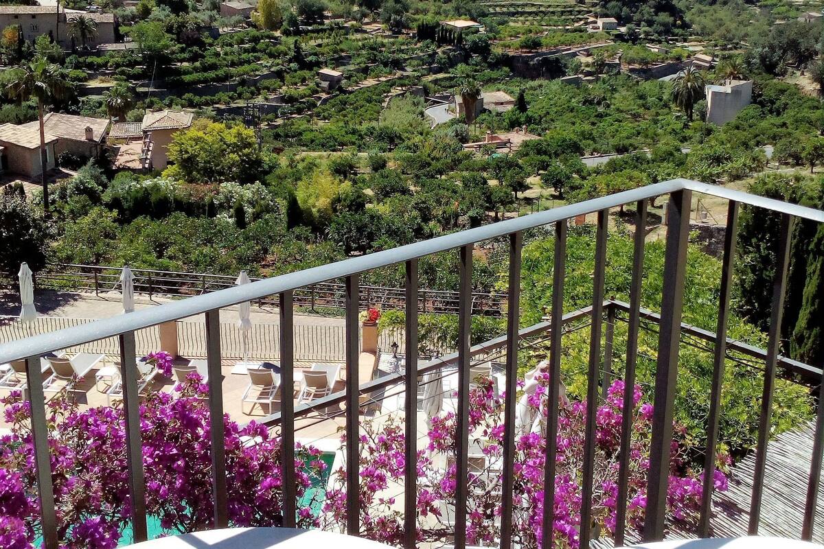 A balcony with white chairs and purple flowers, overlooking a mountainous landscape with terraced fields and buildings.