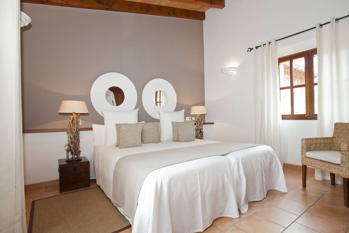 A bedroom with a large bed, two bedside tables with lamps, a wicker chair, and a window with curtains.