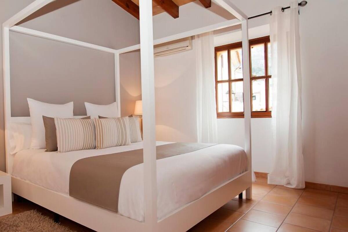 A four-poster canopy bed with white bedding and striped pillows, a bedside table with a lamp, and a window with curtains.