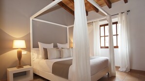Una cama con dosel y cuatro columnas, una mesita de noche con lámpara, un ventanal con cortinas y suelo de baldosas.