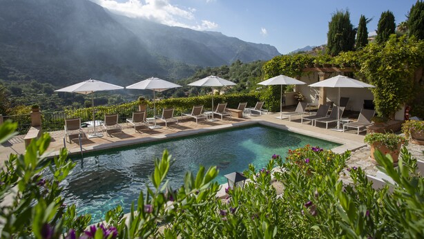 Zona de piscina con sillas de descanso y sombrillas, rodeada de vegetación y montañas al fondo.