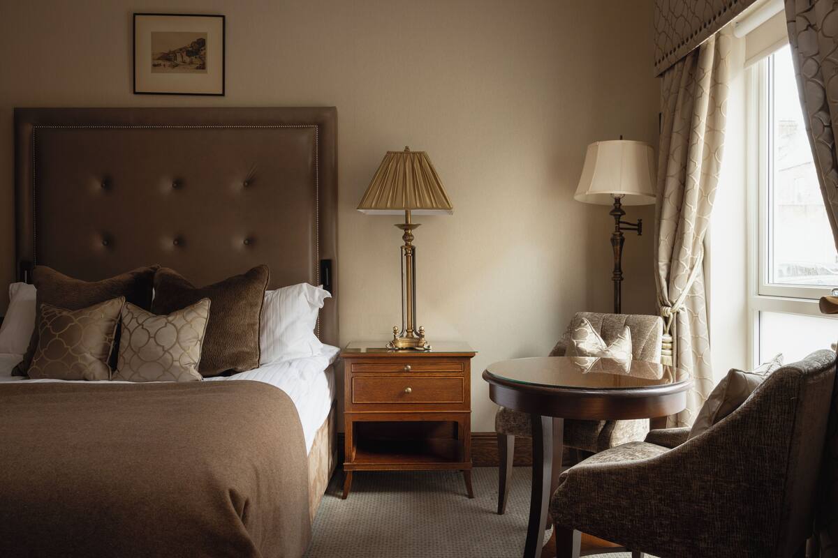 A hotel room with a large bed, two bedside tables, a lamp, a chair, and a window with curtains.