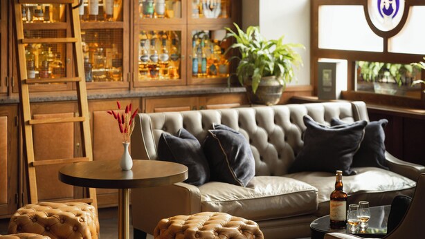 A cozy living room with a leather sofa, matching ottomans, a wooden ladder, and a cabinet filled with bottles. - The Station Hotel (Aberlour)