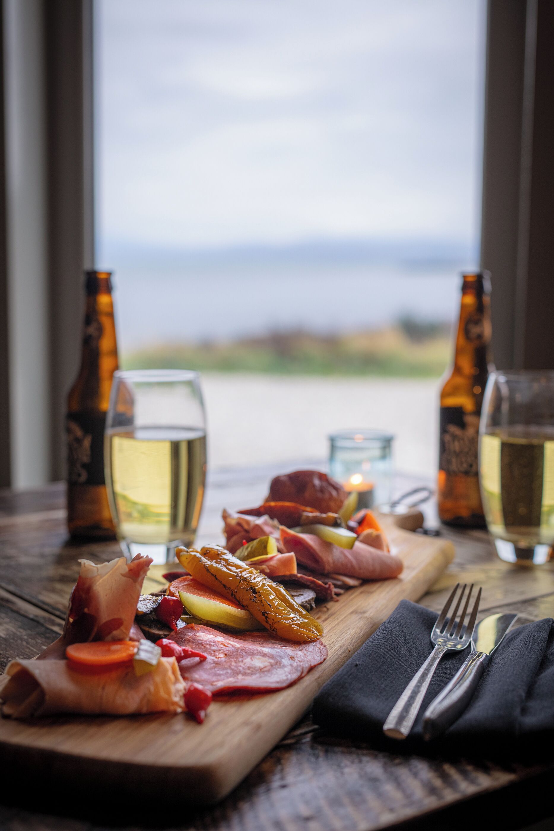 A wooden board with various meats and fruits, two bottles of beer, and glasses of white wine on a table with a view of the outdoors.
