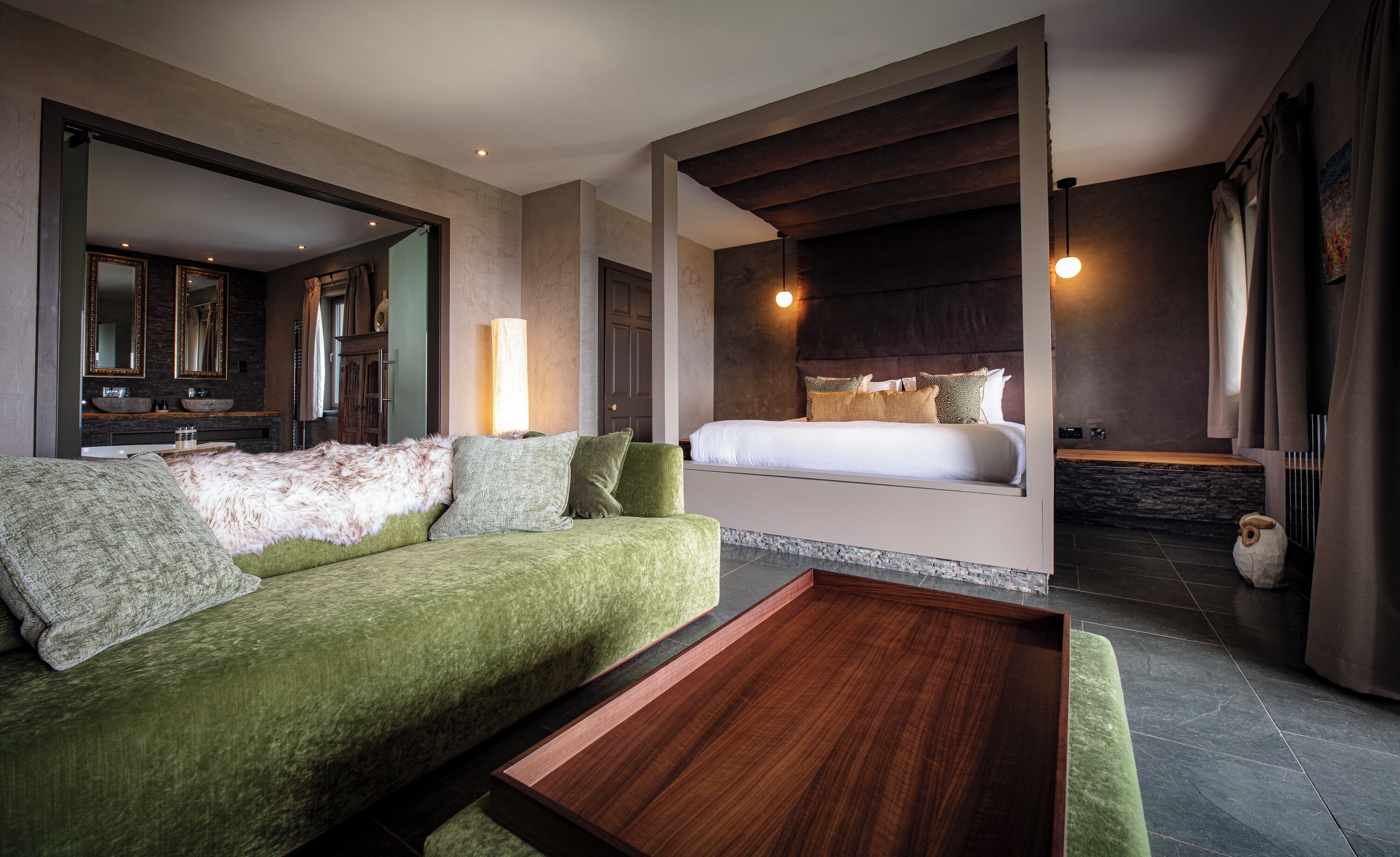 A modern hotel room with a green sofa, a wooden coffee table, a large bed with a headboard, a bathroom with a bathtub, and a large mirror.