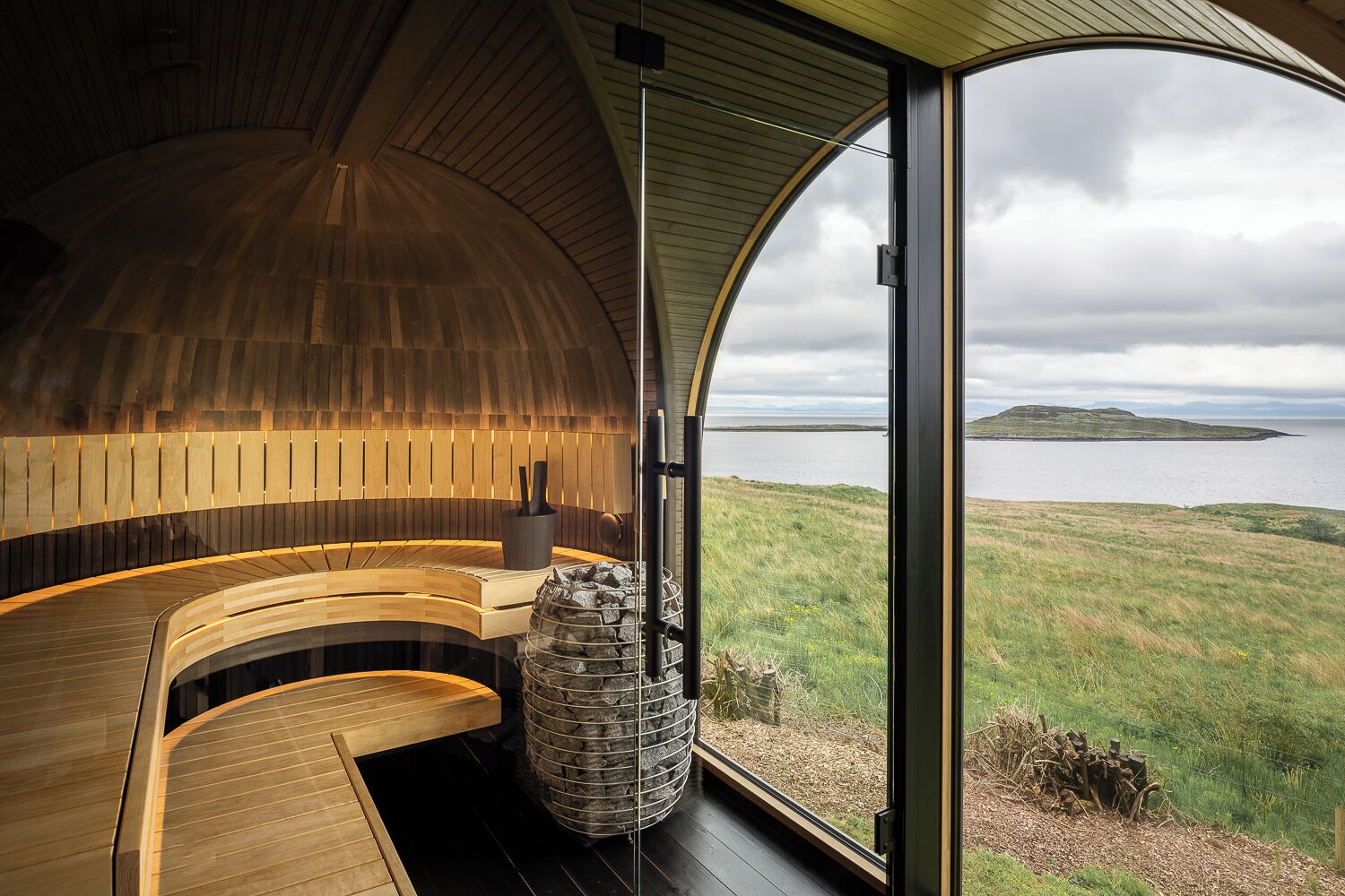 A modern interior with a curved wooden bench and a stone fireplace, overlooking a grassy landscape and water body.