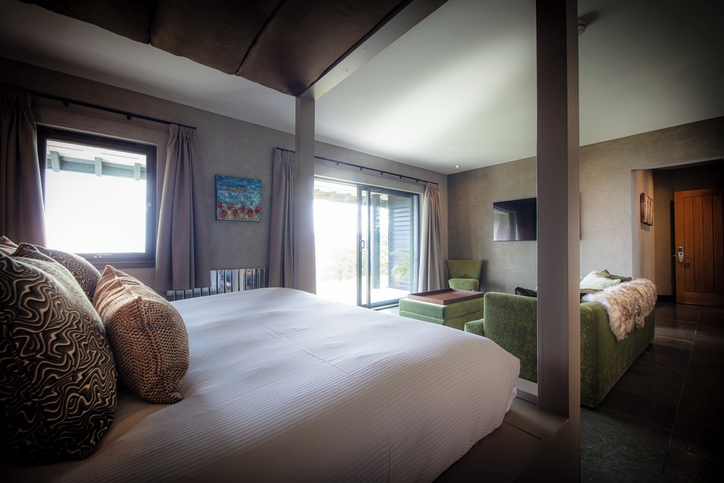 Habitación de hotel con una cama grande, un sillón verde, una mesita pequeña y vistas al exterior a través de una puerta corrediza.