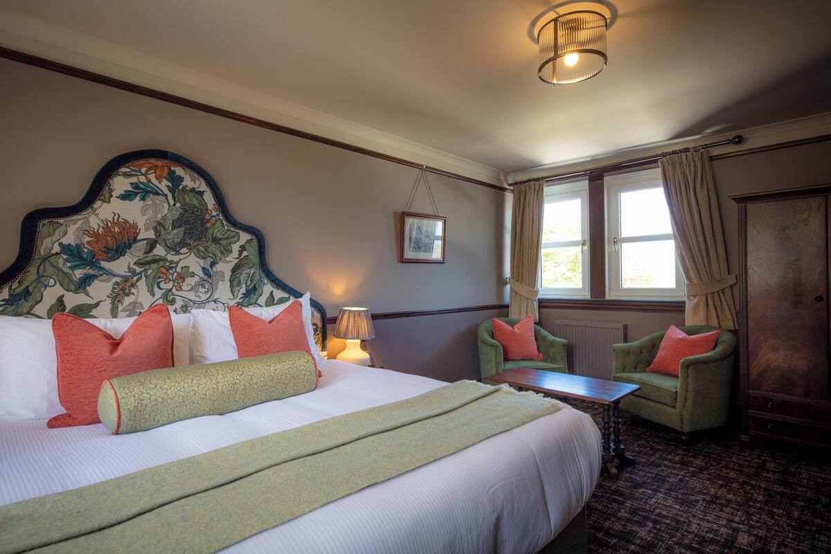A hotel room with a large bed, two armchairs, a small table, and a window with curtains.