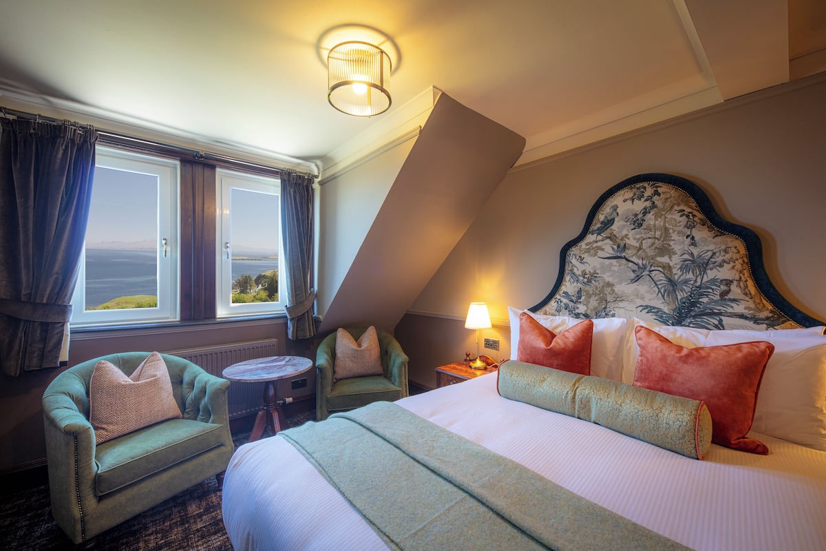 A hotel room with a large bed, a sitting area with a chair and a small table, a lamp, and a view of the sea through the windows.
