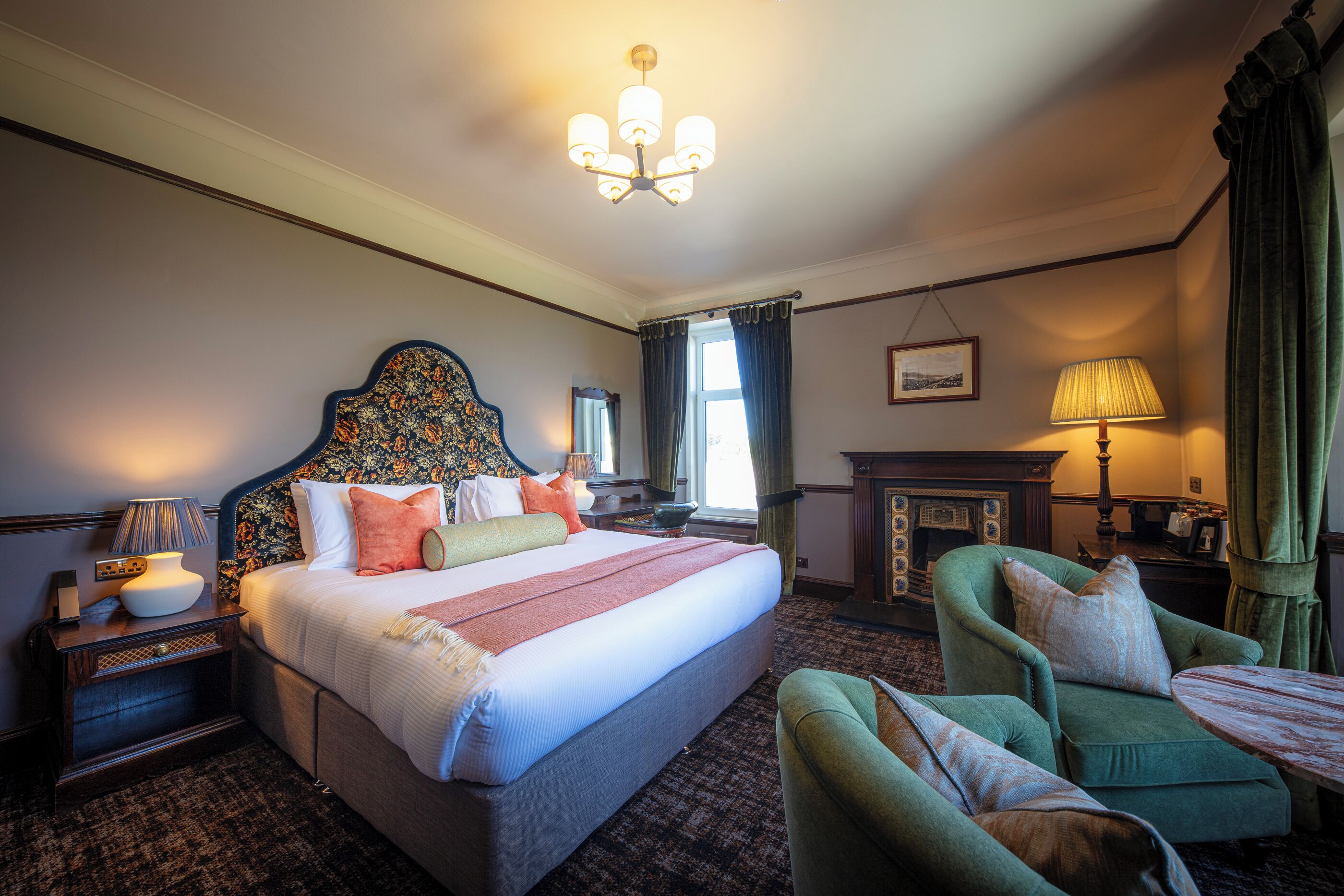 A hotel room with a large bed, a green armchair, a small table, a nightstand, a lamp, a fireplace, and a chandelier.