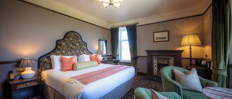 A hotel room with a large bed, a green armchair, a small table, a nightstand, a lamp, a fireplace, and a chandelier.