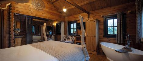 A log cabin bedroom with a large bed, a wooden ceiling, a bathtub, and a window.