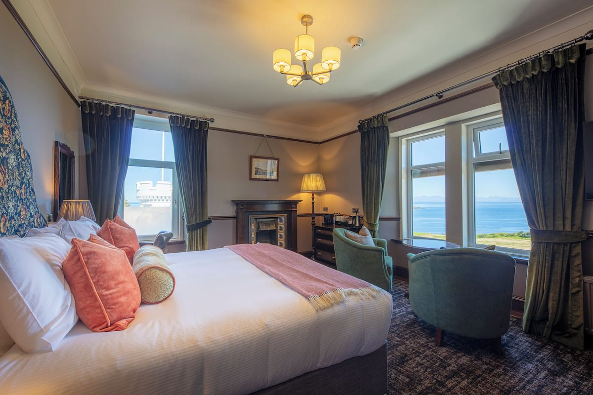A hotel room with a large bed, a green armchair, a fireplace, and a view of the sea.