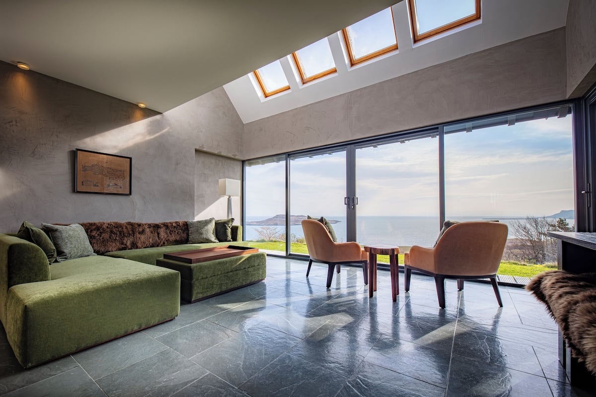 A modern living room with a green sofa, two armchairs, and a view of the sea through large windows.