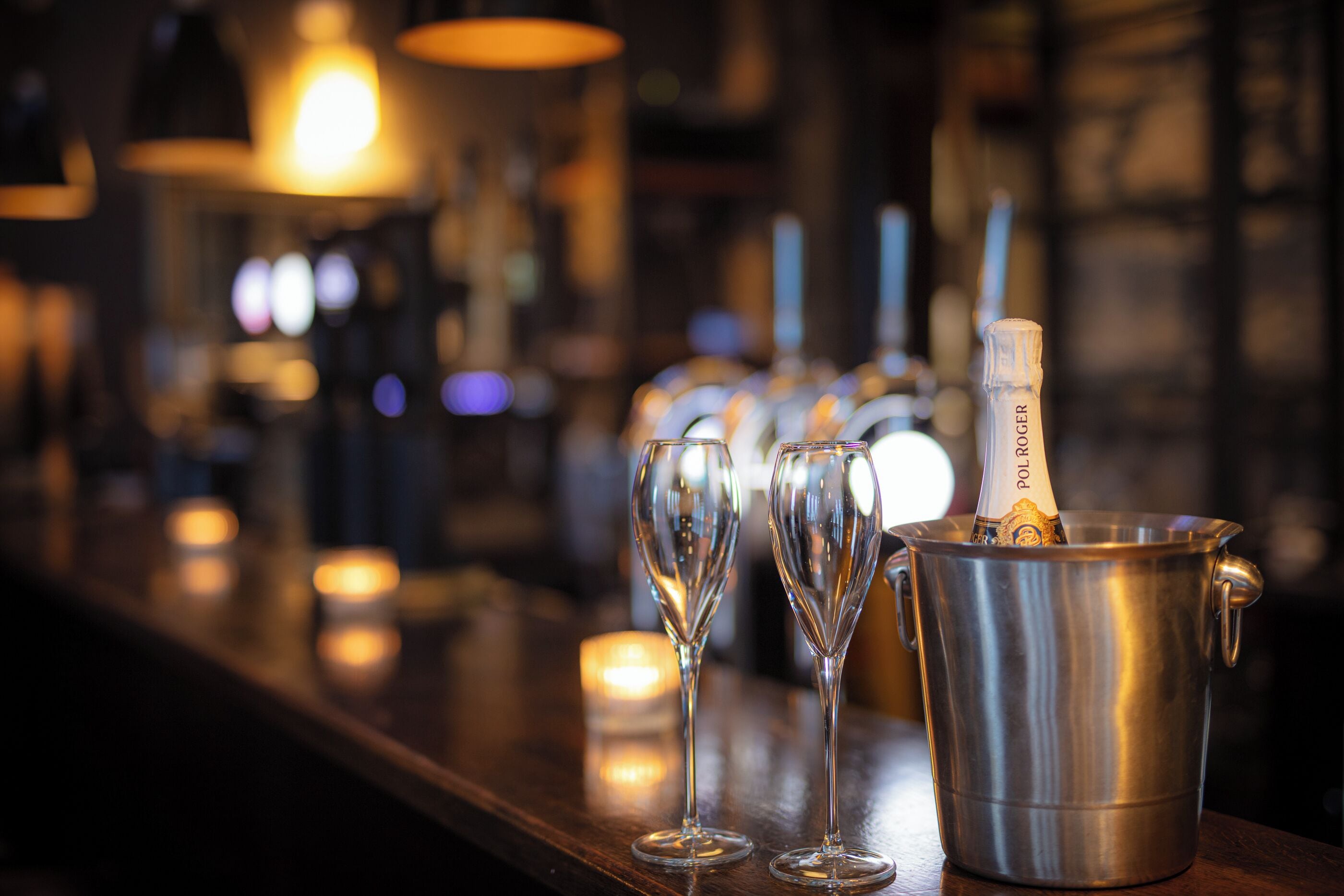 A bar area with a metal bucket containing a bottle of champagne, several empty glasses, and candles in the background.