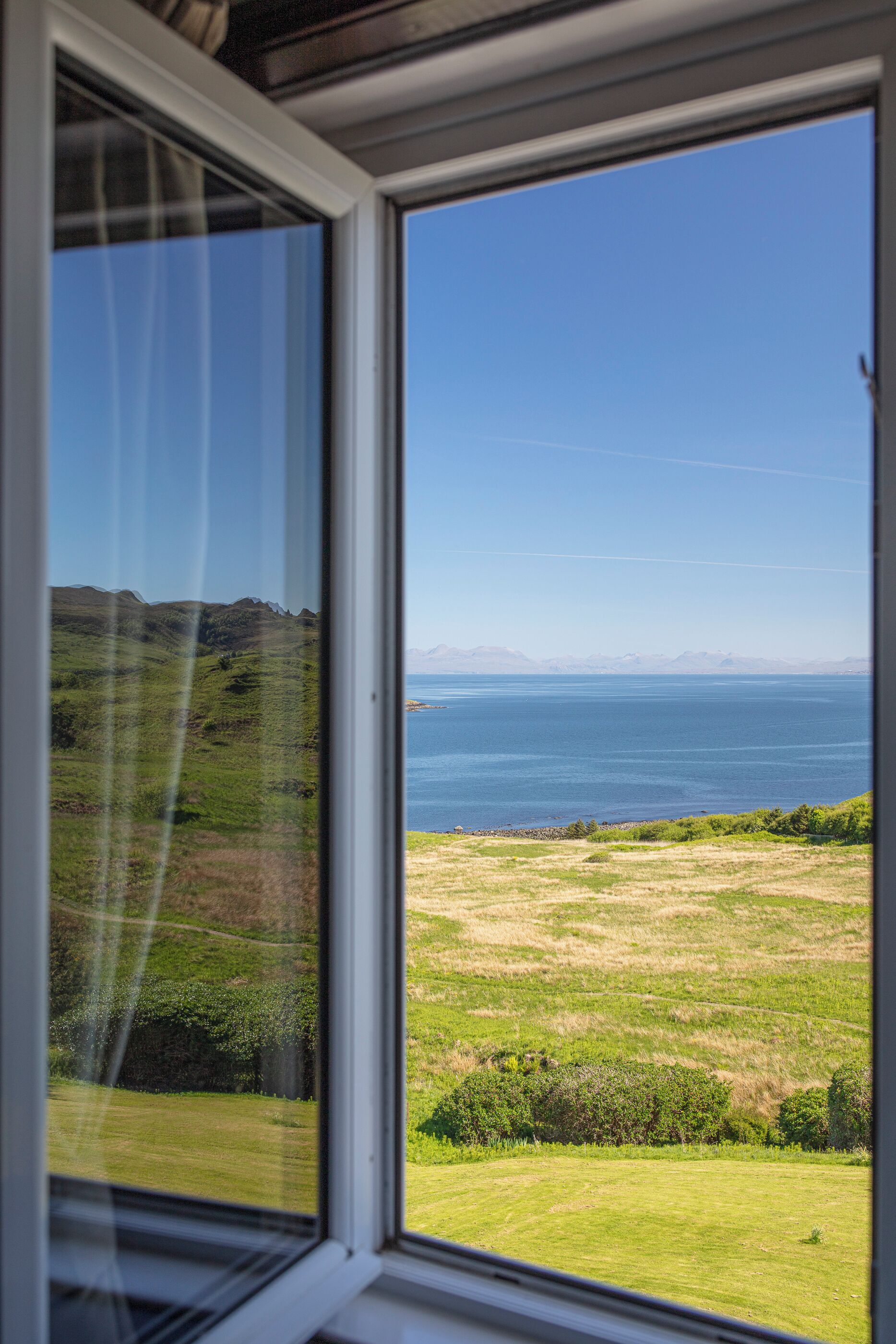 Vista a través de una ventana que muestra un paisaje con praderas, una masa de agua y montañas a lo lejos.