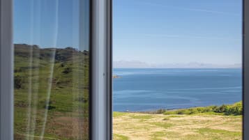 Vista a través de una ventana que muestra un paisaje con praderas, una masa de agua y montañas a lo lejos.