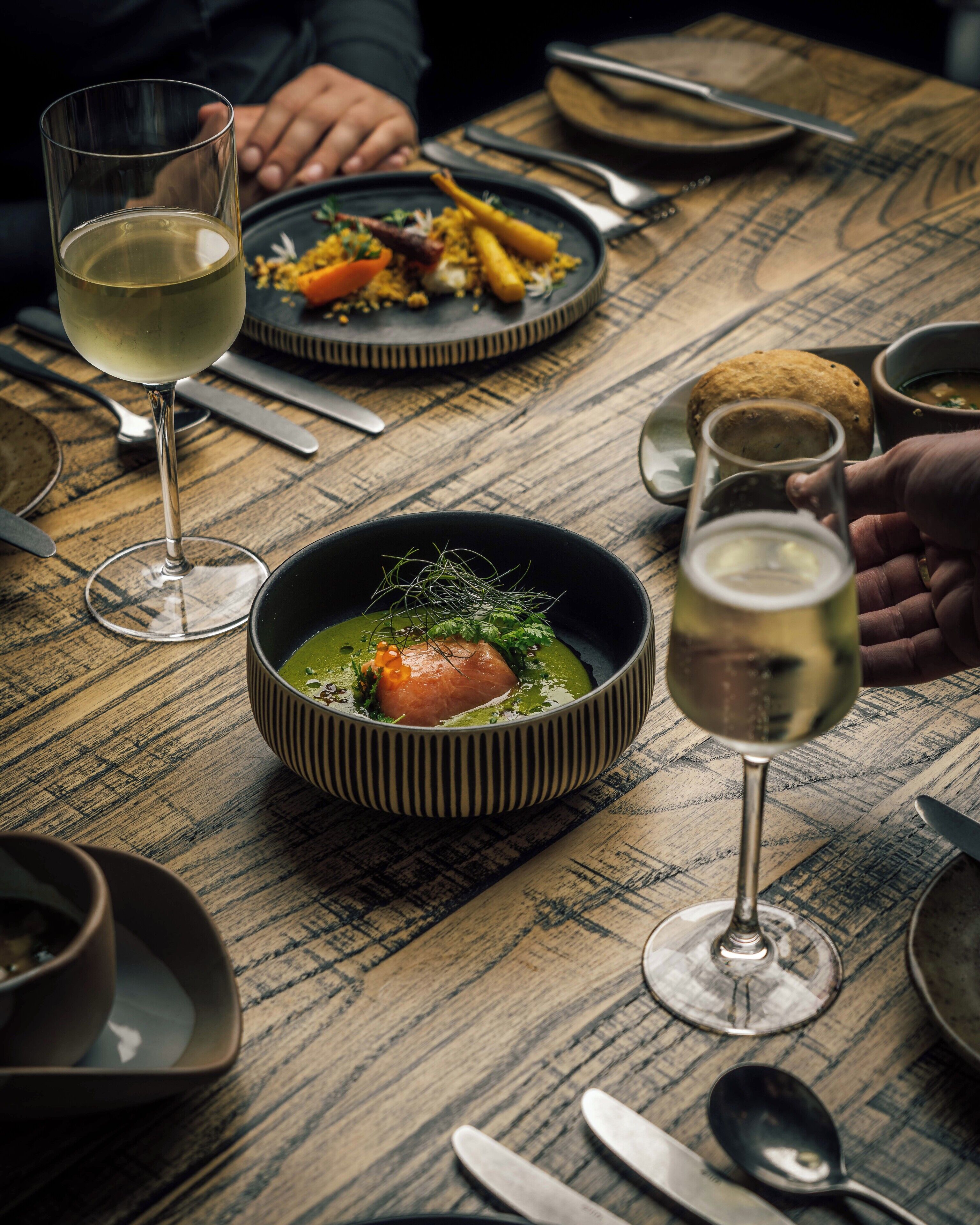 Uma mesa de jantar com uma tigela de sopa, um copo de vinho branco e diversos pratos.