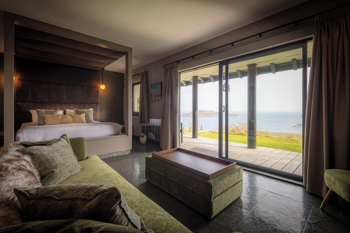 A modern bedroom with a large bed, a green sofa, and a view of the sea through large sliding doors.