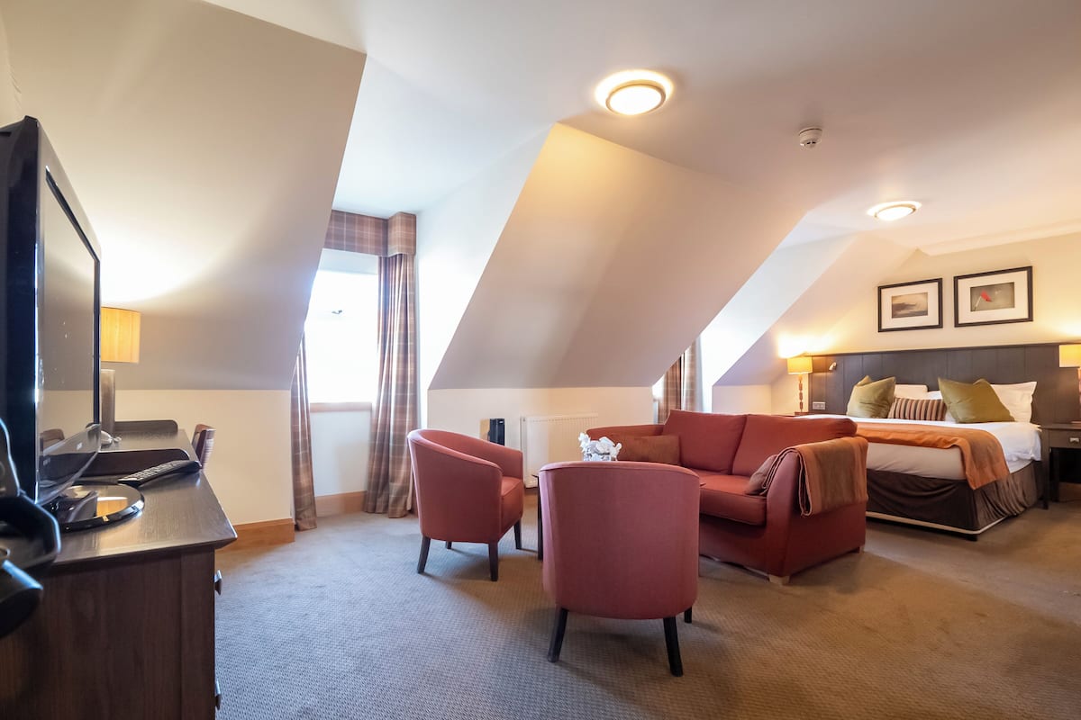 A hotel room with a large bed, a sofa, two armchairs, a desk with a computer, and a television.