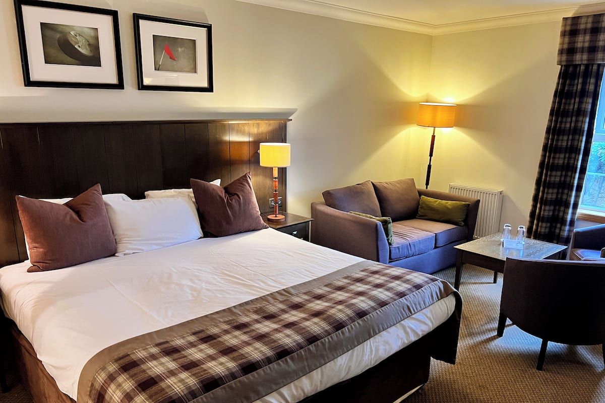 A hotel room with a large bed, a sofa, a small table, and a lamp. There are framed pictures on the wall and a window with curtains.